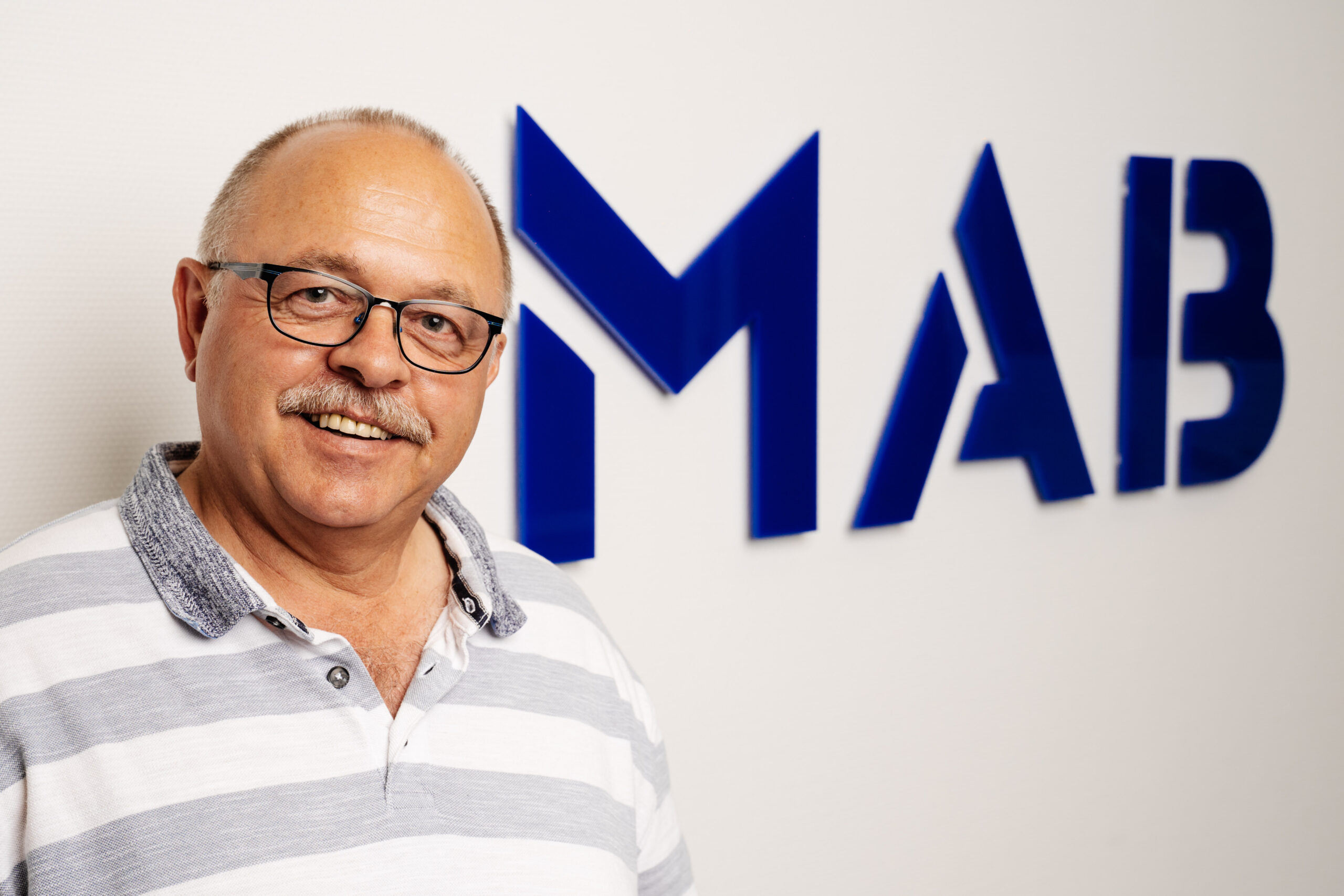 Mann mit Brille und Schnurrbart lächelt vor blauem Firmenlogo an weißer Wand